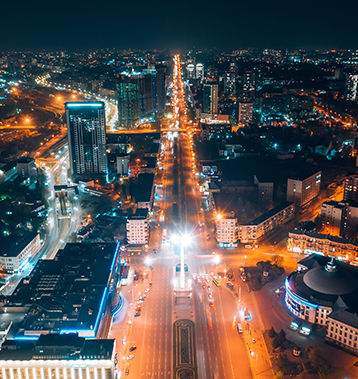 Vista aérea noturna de uma cidade iluminada, com avenidas movimentadas em que as luzes dos carros formam rastros, prédios com luzes de escritórios acesas e um calçadão central destacando-se na paisagem urbana.